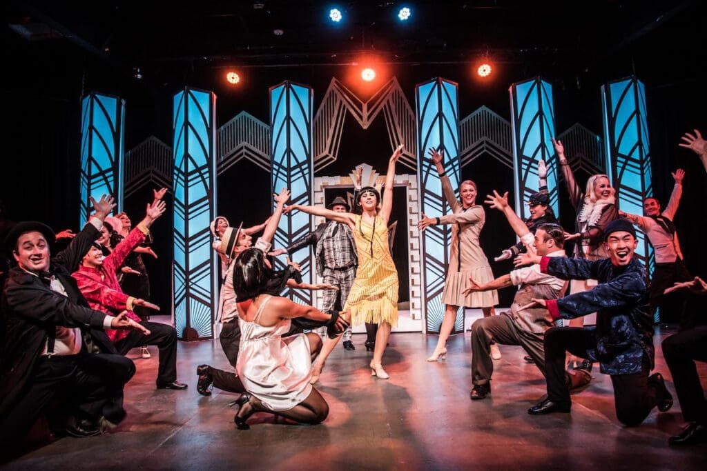 A vibrant theater performance featuring a group of actors in 1920s-style costumes. A woman in a yellow flapper dress dances at the center, surrounded by lively cast members in dynamic poses against an Art Deco backdrop.