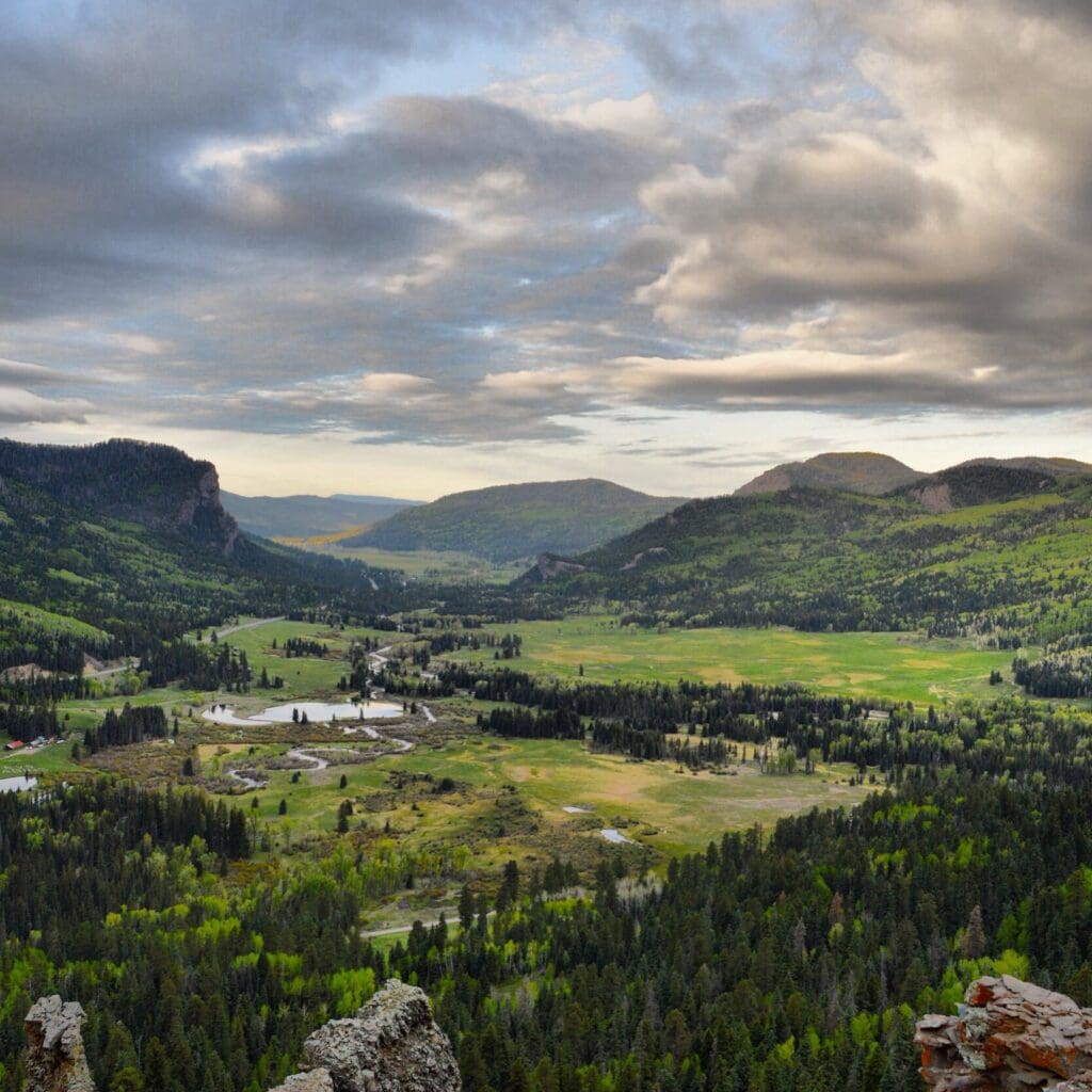 A scenic view of a lush green valley surrounded by rolling hills under a partly cloudy sky. The landscape includes patches of forest, open meadows, and a river winding through the valley. Hills are lightly shadowed under scattered clouds.