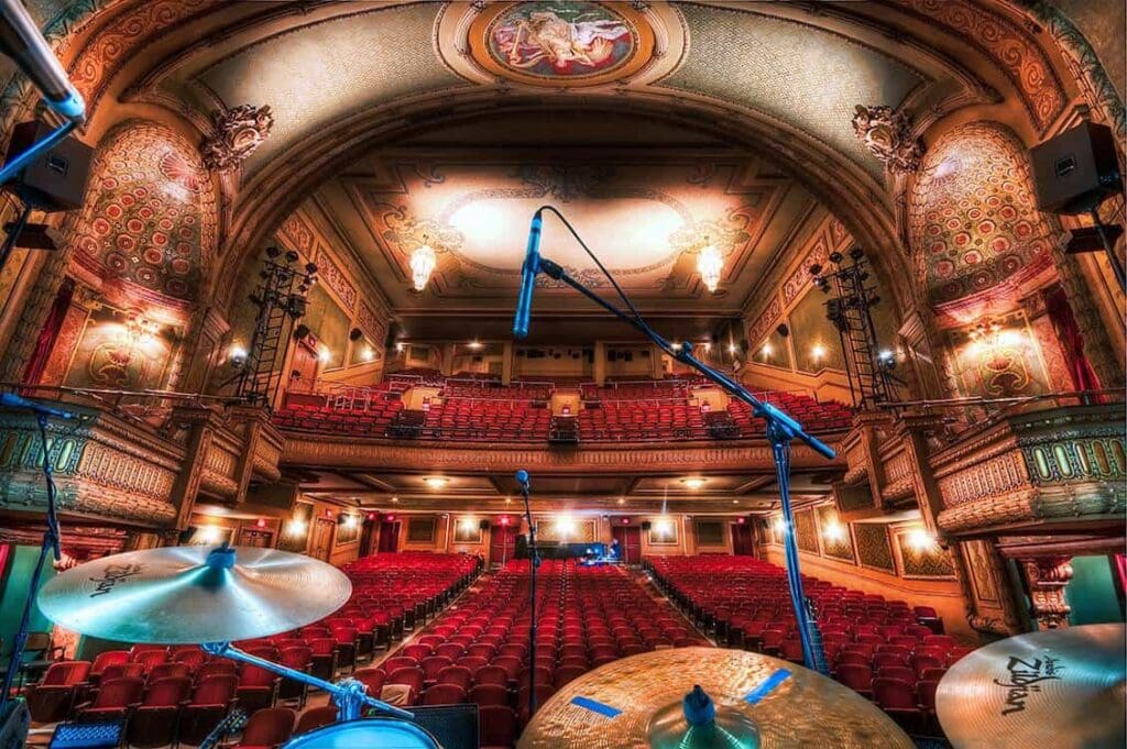 View from a theater stage with a drum set and microphones in the foreground. The ornate auditorium features rows of red seats, decorative walls, and a large, illuminated ceiling design, suggesting an opulent atmosphere.