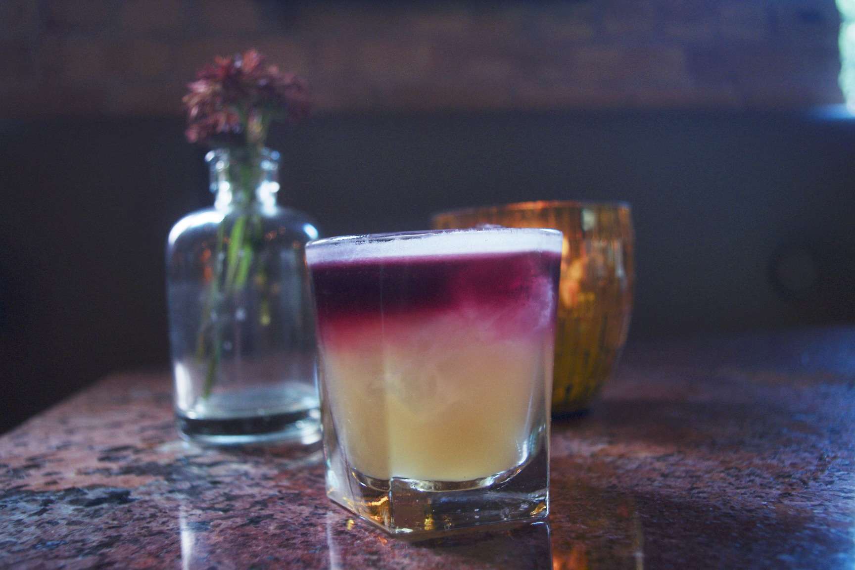 A cocktail in a short glass with a yellow base and red top sits on a table. A glass vase with dried flowers and a lit amber candle are blurred in the background.