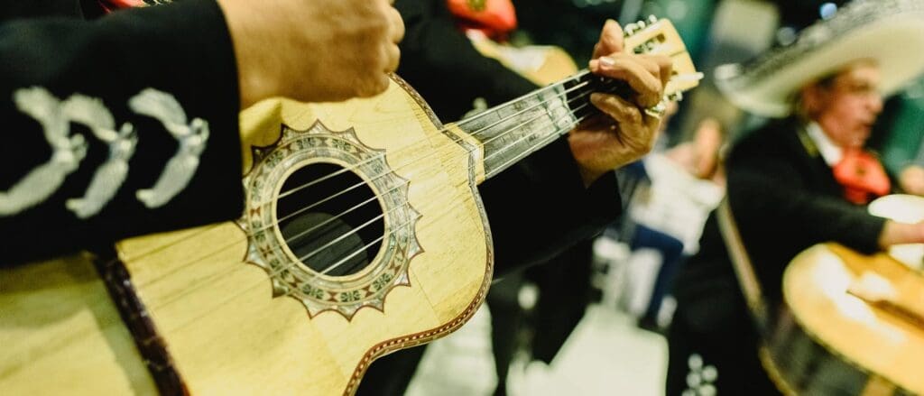A close-up of a musician playing a guitar-like instrument, part of a mariachi band. The focus is on the intricate design of the instrument, with blurred figures in traditional mariachi attire in the background.