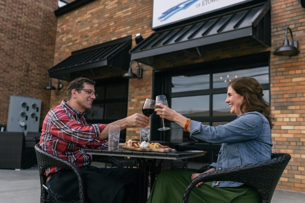 A man and woman sit at an outdoor table clinking wine glasses. The man wears a red plaid shirt and glasses, and the woman has a denim jacket and brown hair. They are smiling with a meal on the table, in front of a brick building.