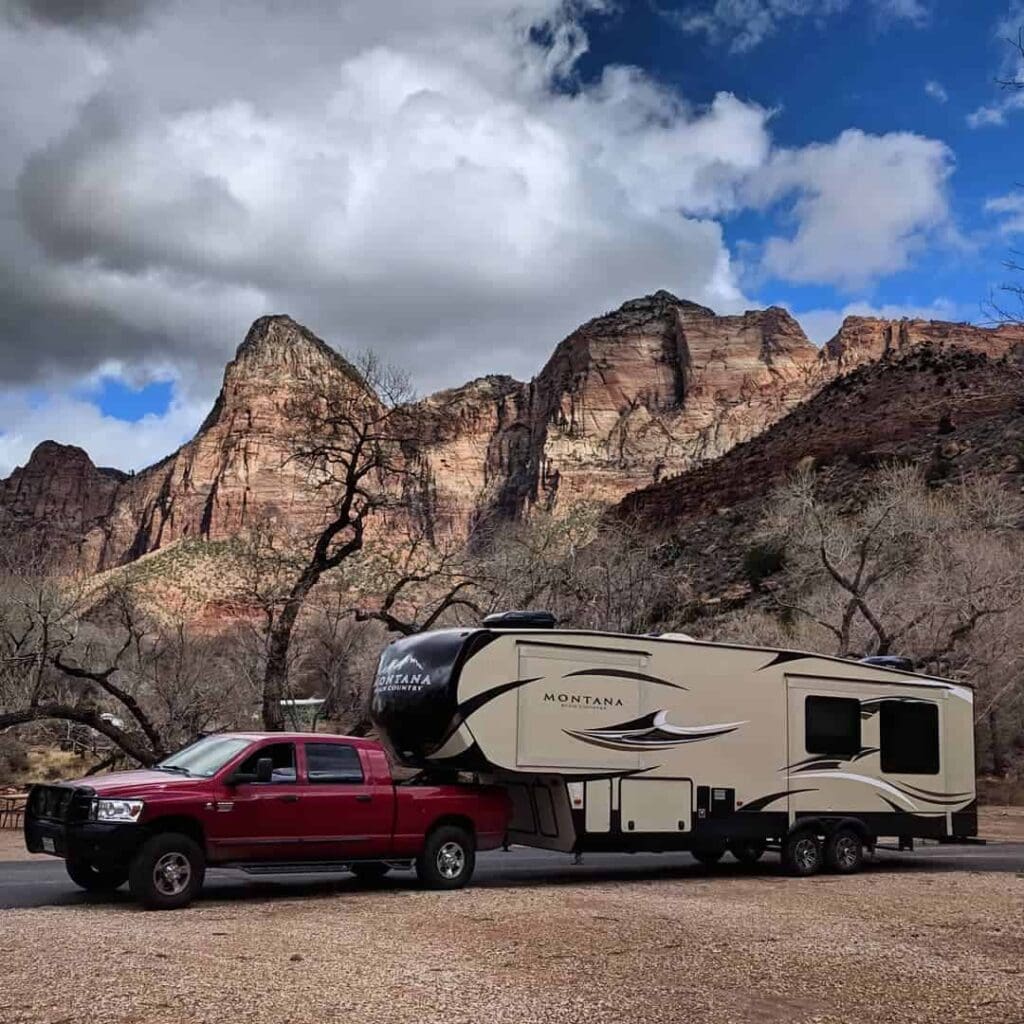 A red truck is towing a beige Montana RV trailer against the backdrop of dramatic mountains and cloudy skies. Bare trees are visible in the foreground, suggesting a wintry setting.