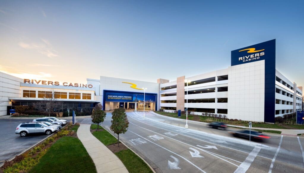 Wide-angle view of Rivers Casino entrance and parking garage. The building is modern with blue and white signage. Some cars are parked, and others are driving on the road in the foreground. The sky is clear, suggesting its daytime.
