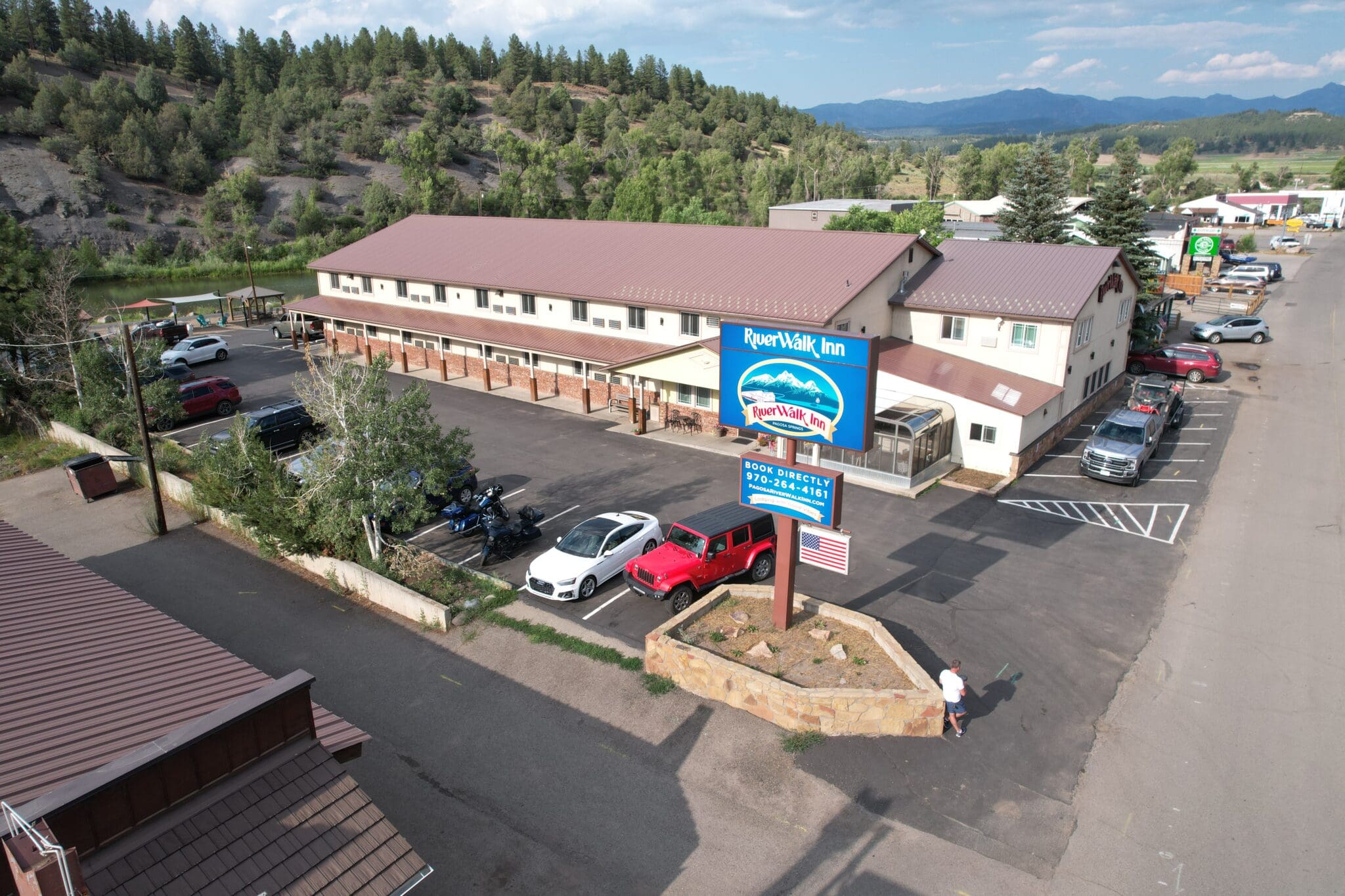 Aerial view of a two-story motel with a prominent RiverWalk Inn sign. The parking lot has several cars and a person walking. Surrounded by trees and mountains, with a blue sky overhead.