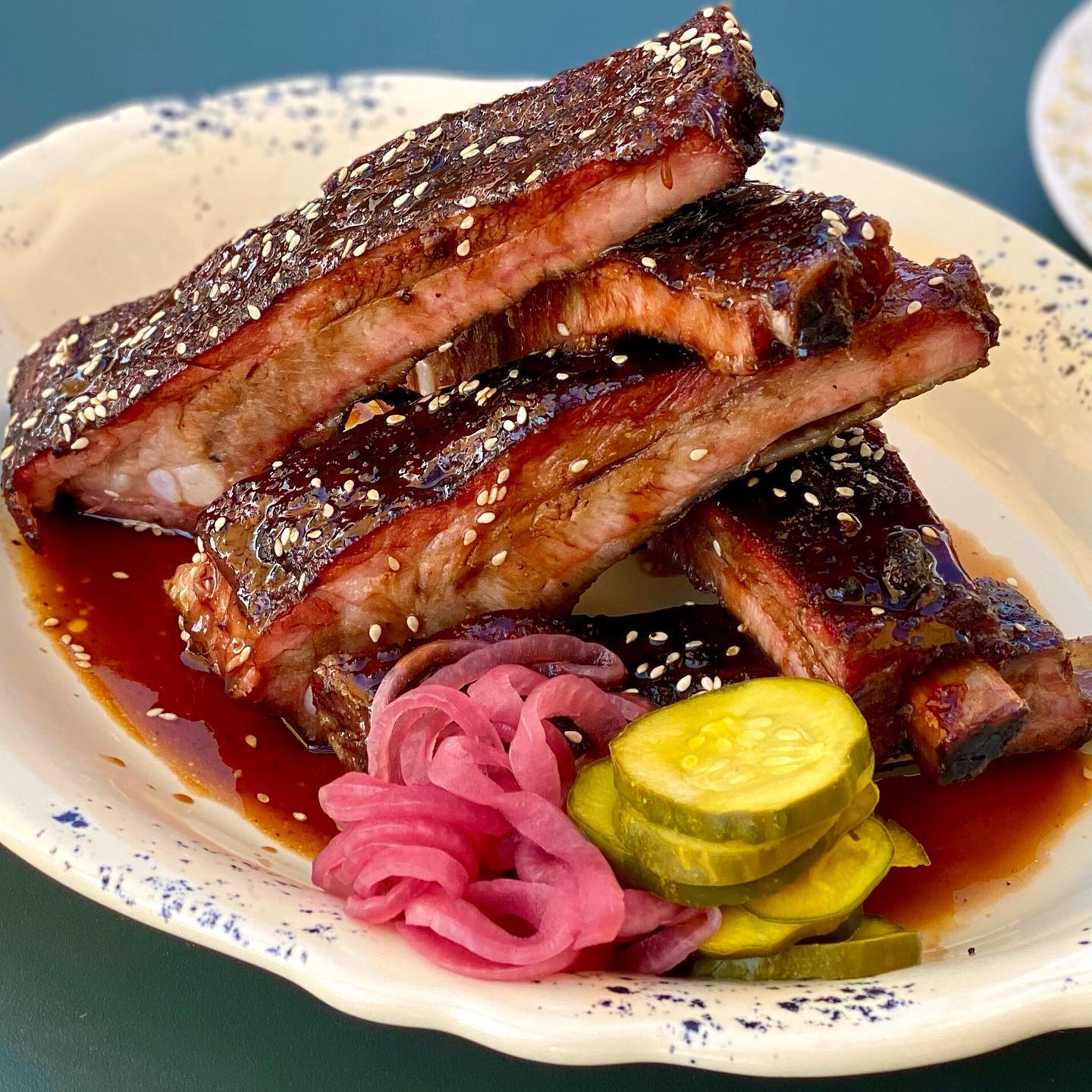 A plate of barbecued ribs drizzled with sauce and sprinkled with sesame seeds. Accompanied by pickled red onions and sliced pickles. The dish is served on a speckled white plate with a teal background.