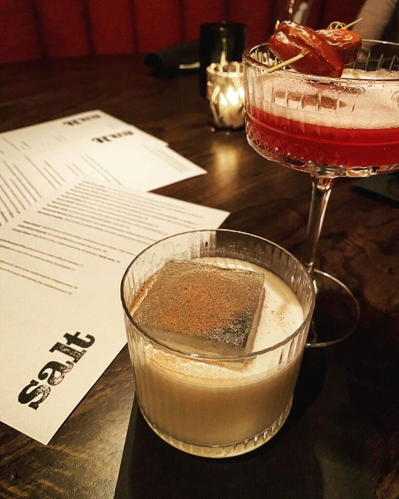 Two cocktails on a table: one with a large ice cube in a short glass, the other red with a garnish in a coupe glass. A menu with salt in bold letters is beside them, and a small candle glows softly in the background.