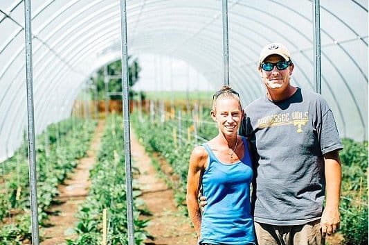 A woman and a man stand smiling inside a greenhouse. They are surrounded by rows of plants. The woman is wearing a blue tank top, and the man is wearing a cap and t-shirt. The greenhouse structure is visible overhead.