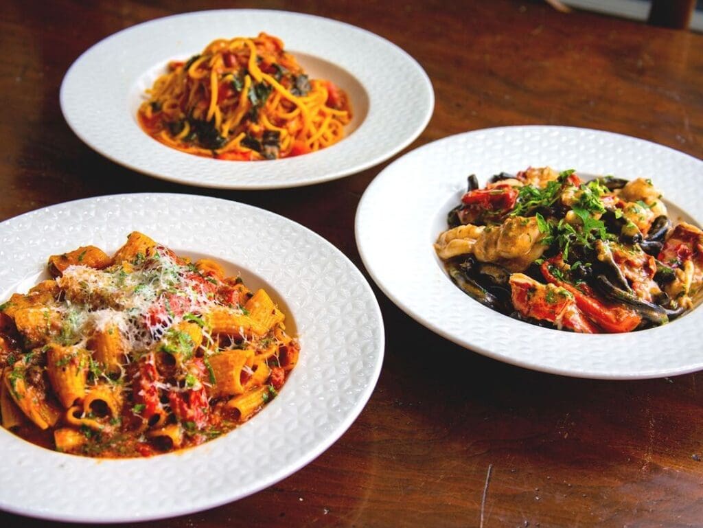 Three plates of pasta are on a wooden table. The first plate has spaghetti with tomato sauce and herbs. The second has rigatoni with grated cheese and herbs. The third features pasta with seafood and vegetables, garnished with parsley.