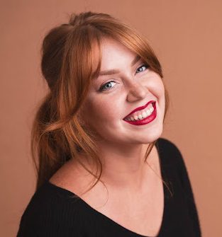 Smiling woman with long red hair and bright red lipstick, wearing a black top, against a warm, neutral background.