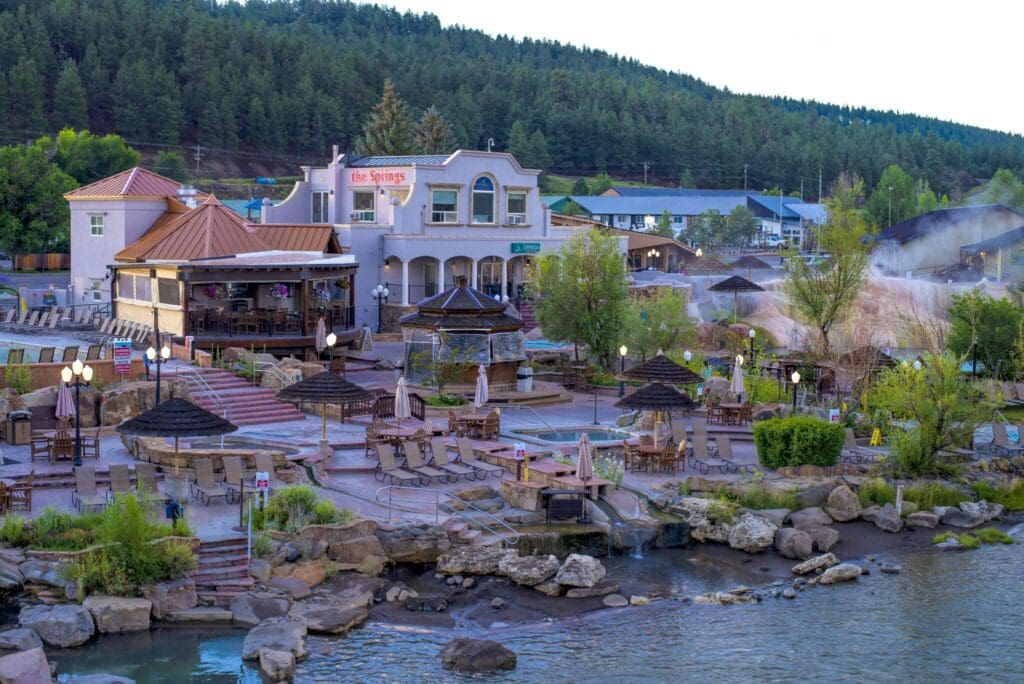Outdoor hot springs resort with pools and seating areas surrounded by landscaped greenery. A multi-story building labeled the Springs sits amidst a natural setting with trees and a river. Steaming water is visible near the pools.
