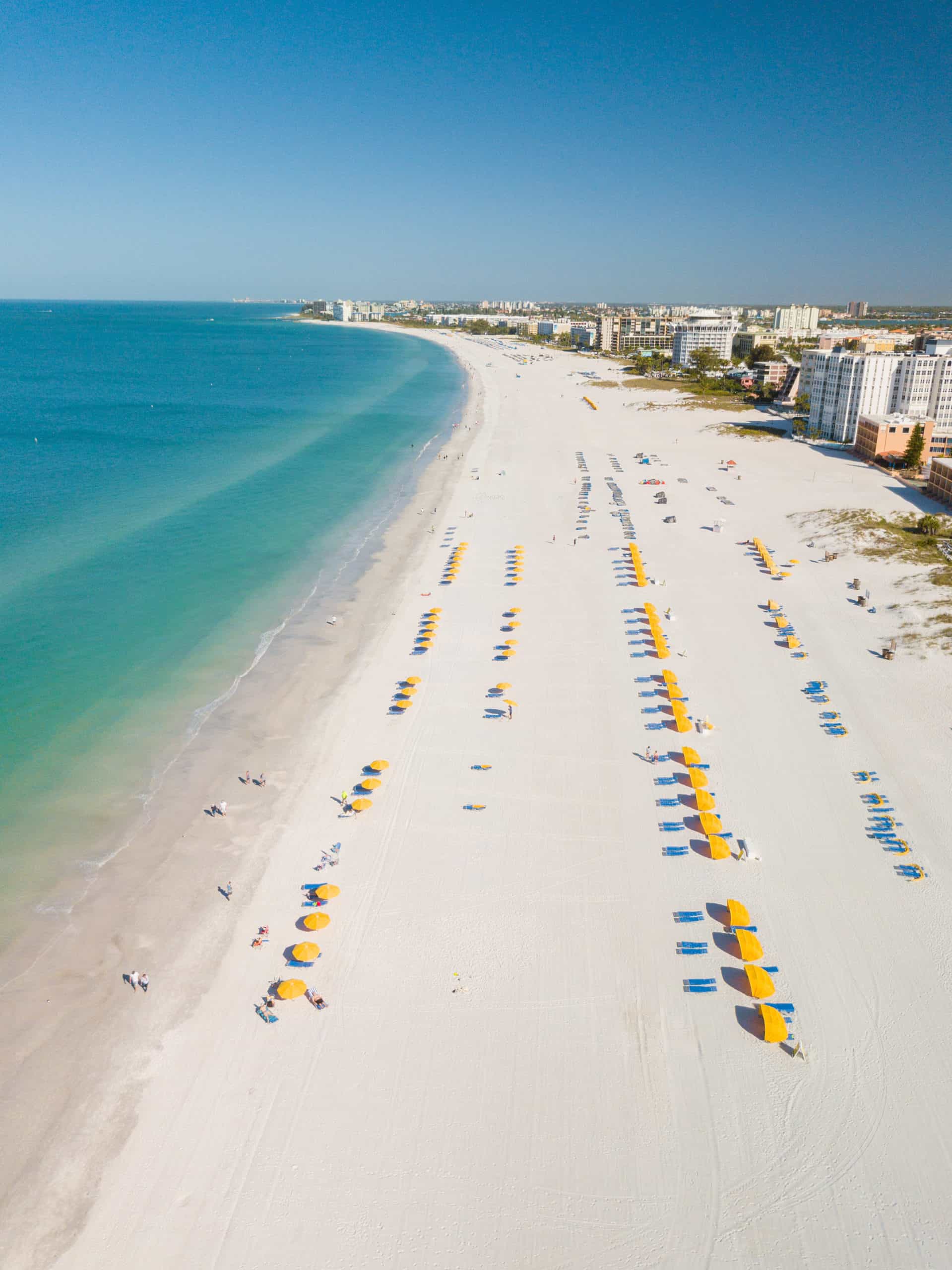 Aerial view of a sandy beach with rows of yellow and blue umbrellas, adjacent to a turquoise ocean. Buildings line the shore in the background under a clear blue sky, with people scattered across the beach.