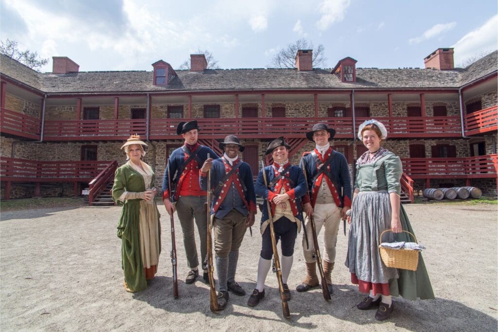 A group of six people dressed in colonial-era clothing stand in a courtyard. Three hold muskets, and one carries a basket. They are in front of a stone building with red wooden accents, possibly part of a historical reenactment setting.