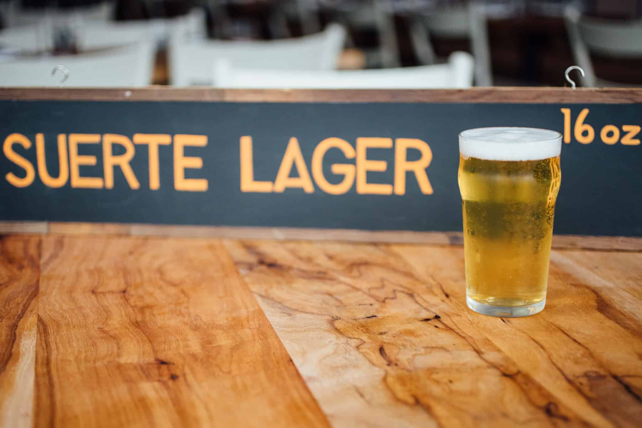 A pint of lager sits on a wooden table, with a blackboard behind it displaying Suerte Lager 16 oz in yellow text. Tables and chairs are blurred in the background.