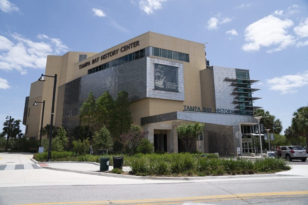 Street view of the Tampa Bay History Center, a modern beige and gray building with large windows. The name is prominently displayed on the facade. The scene includes blue skies, scattered clouds, and surrounding greenery.