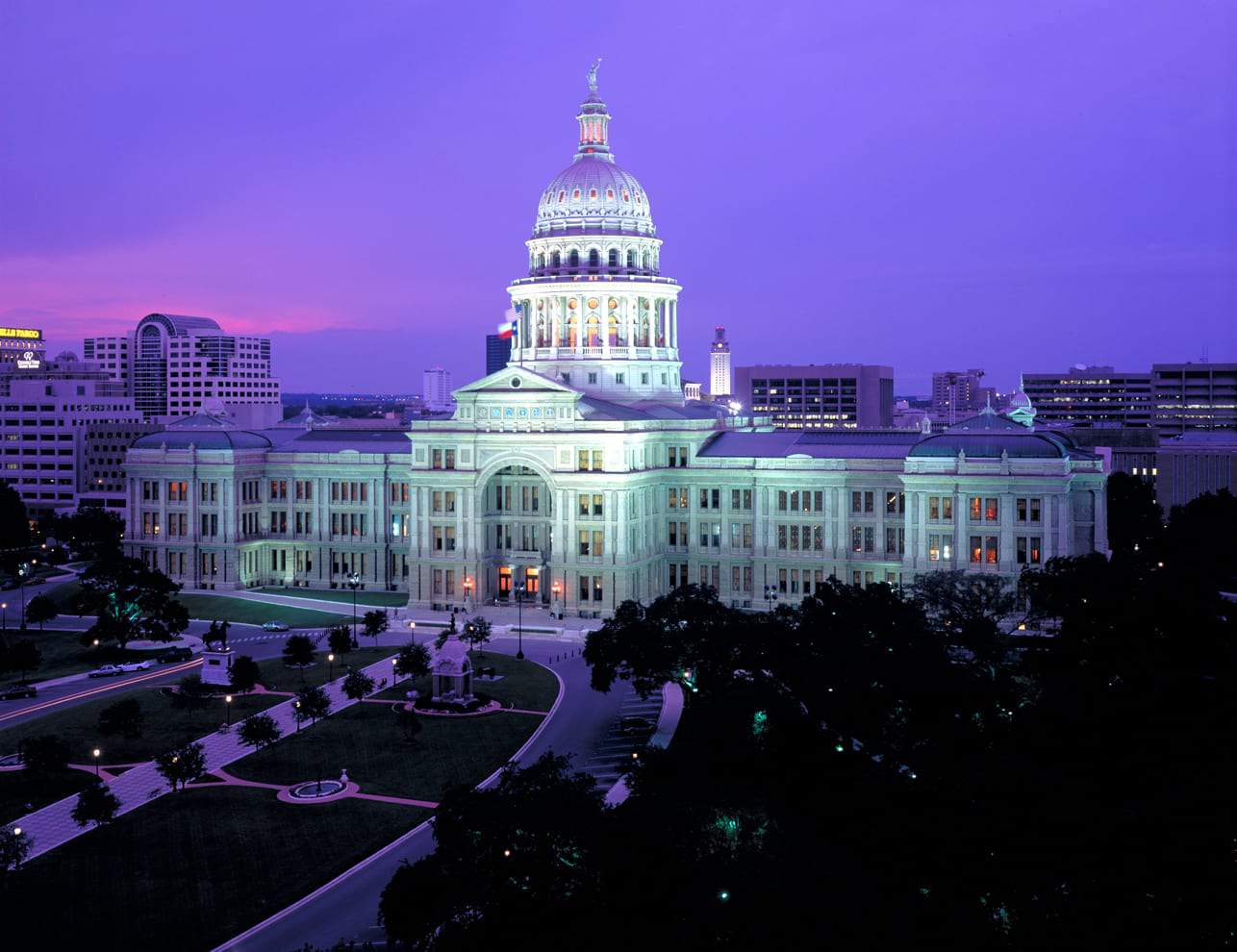 A grand domed building lit up at night, surrounded by pathways and trees. The sky is a deep purple, suggesting twilight. The architecture is classical with a prominent dome and detailed facade. City buildings are visible in the background.