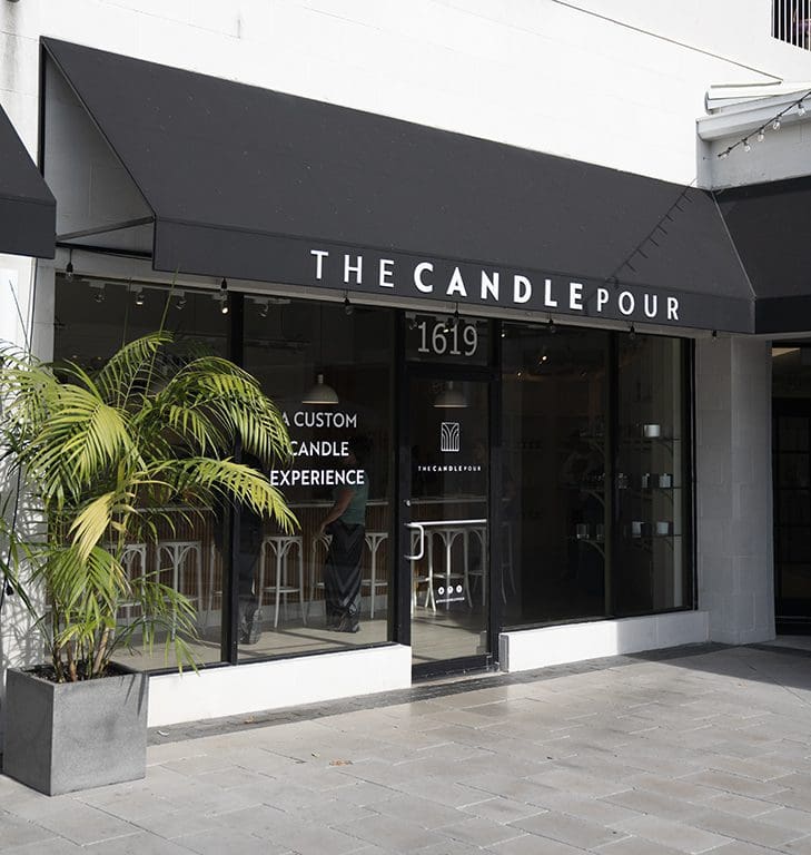 Storefront of The Candle Pour with a black awning and large glass windows. The entrance is flanked by a potted plant. The sign reads A Custom Candle Experience with stools visible inside.