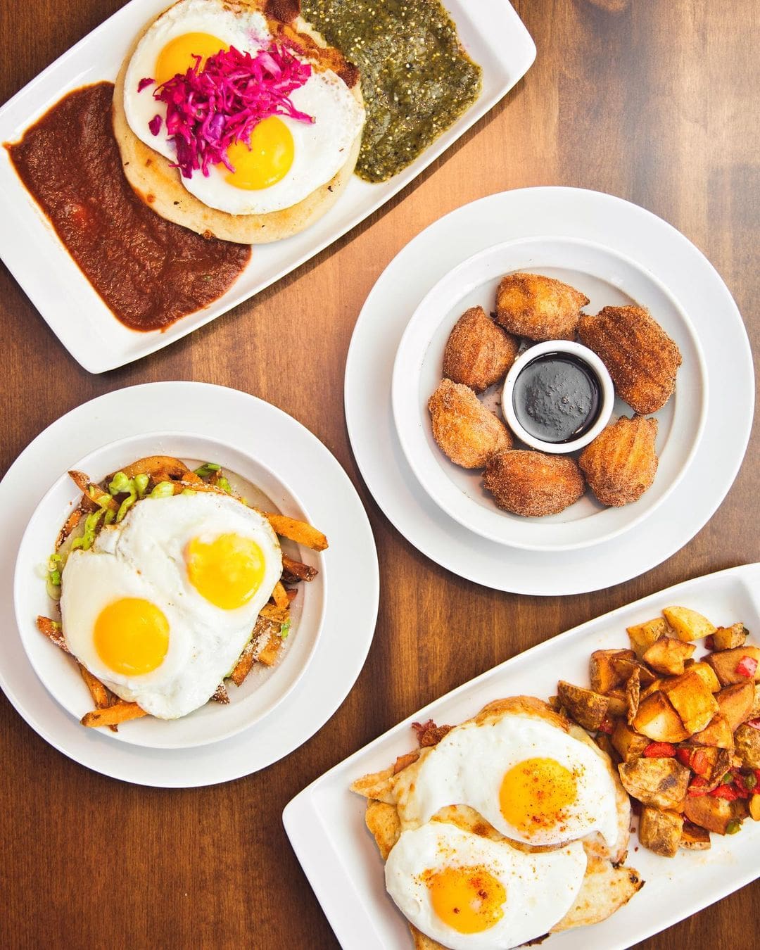Four plates of food are arranged on a wooden table. They include sunny-side-up eggs on tortillas with different sauces, churros with dipping sauce, and hash browns. Each dish is garnished with a variety of toppings and sides.