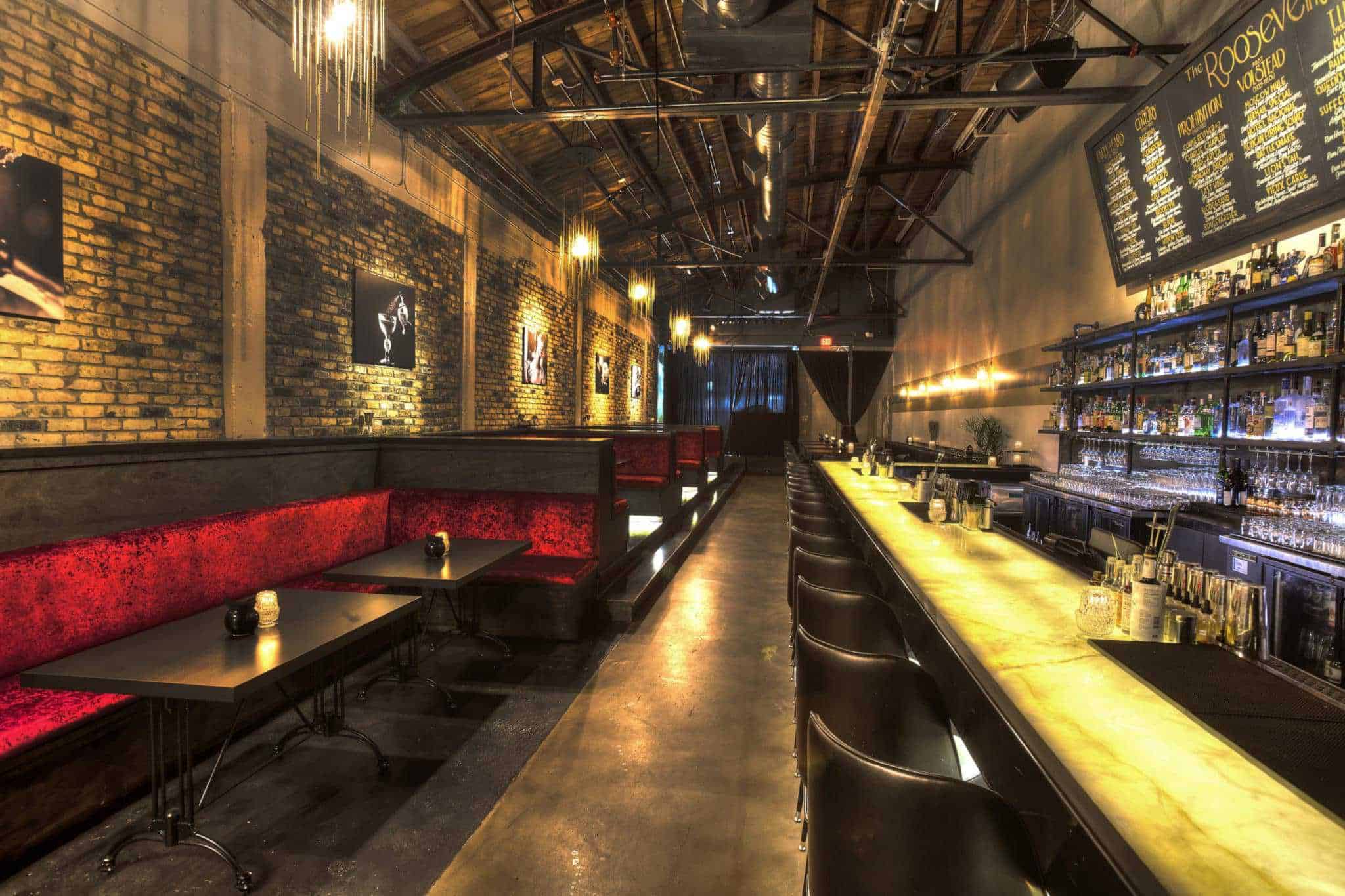 A dimly lit bar with a long counter on the right featuring bottles and glasses. Red velvet booths line the left side, with exposed brick walls adorned with framed pictures. Hanging lights and a menu board enhance the cozy atmosphere.