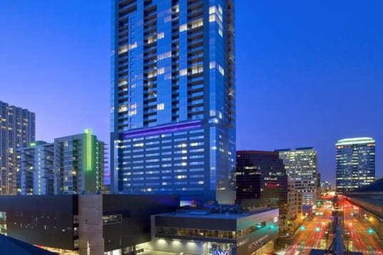 A tall skyscraper stands illuminated against a twilight sky, surrounded by other lit-up city buildings. Streets below are bustling with lights from traffic. The scene captures a vibrant urban atmosphere.