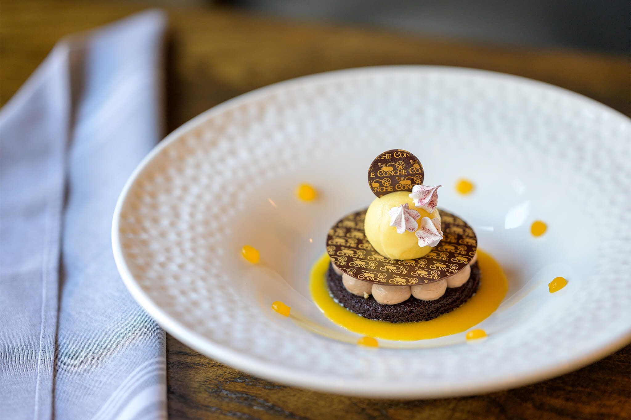 A dessert featuring layers of chocolate and cream on a white plate, topped with a small scoop of yellow ice cream, edible flowers, and a decorative chocolate piece. The plate is garnished with orange sauce dots, placed on a wooden table near a folded napkin.