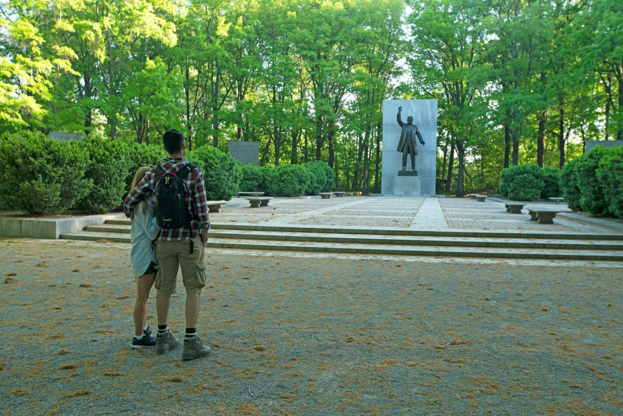 A couple stands on a gravel path in a park, looking at a statue of a person with one arm raised. The area is surrounded by green trees and well-maintained bushes, with sunlight filtering through the foliage.