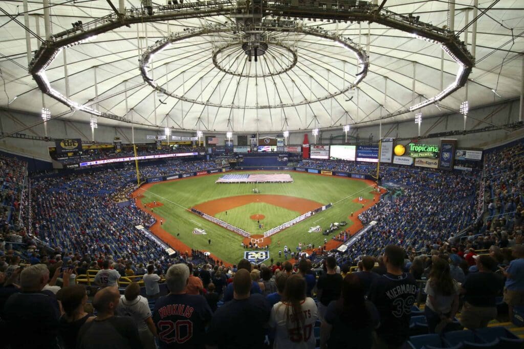 A wide view of a domed baseball stadium filled with fans. The field is prepared for a game, with a large American flag spread across the outfield. Players and officials stand along the baselines. Banners and ads adorn the stadium walls.
