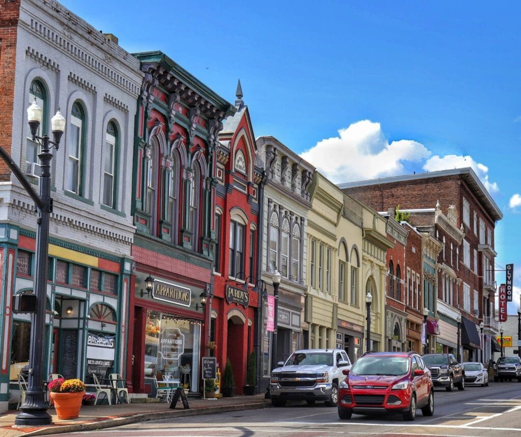 A picturesque street scene with colorful historic buildings, including shops and eateries, under a blue sky. Cars are parked along the street, and a red car is driving by. Lamp posts and a potted plant add to the charming atmosphere.