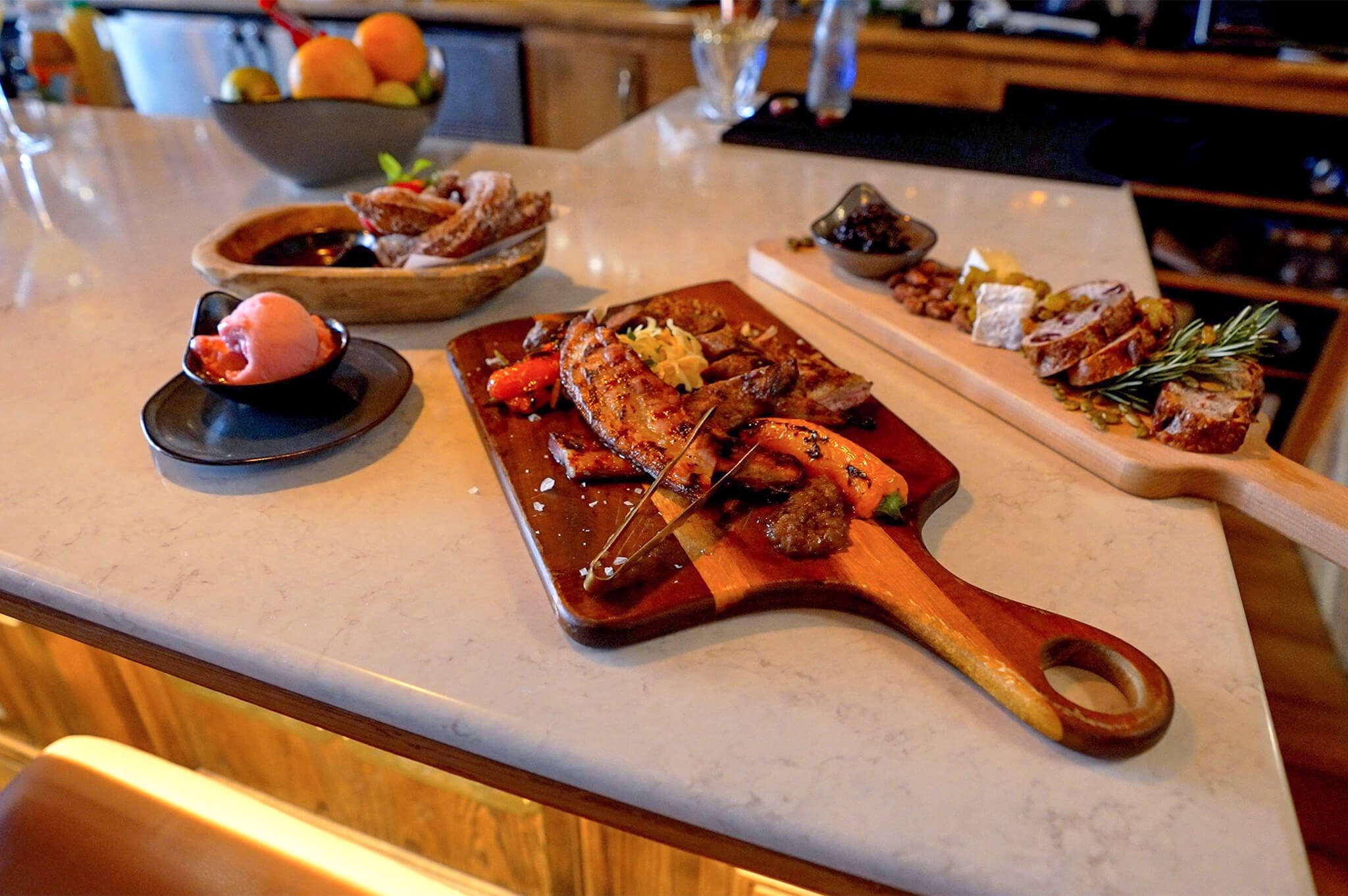 A marble countertop displays a variety of dishes, including a wooden platter with grilled meats and vegetables, a bowl of bread slices, and a small dish with a scoop of red sorbet. A bowl of fruit is in the background.