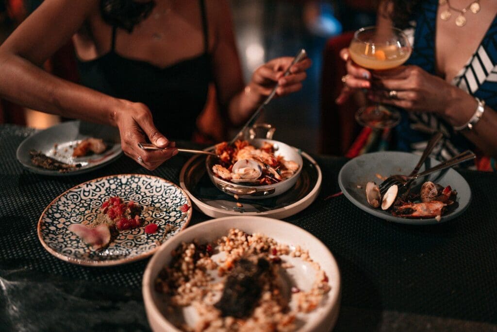 Two people dining with a variety of dishes on the table, including seafood and grains. One person uses chopsticks, while the other holds a cocktail glass. The scene is warmly lit, creating a cozy atmosphere.
