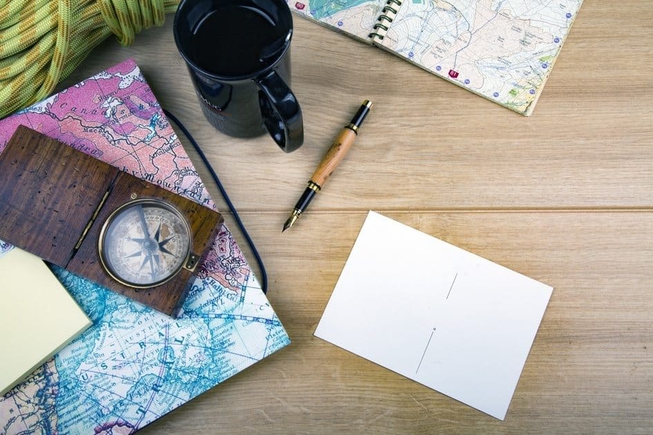 A wooden table displays a compass, maps, a black mug, a notebook, a pen, and a piece of blank paper. The maps are colorful, featuring roads and topographical details. The arrangement suggests planning for a journey or adventure.
