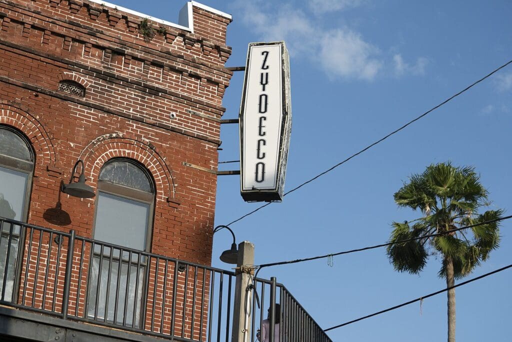 A vintage-style building with red brick and arched windows, featuring a vertical sign that reads Zydeco. A palm tree and power lines are visible under a clear blue sky.