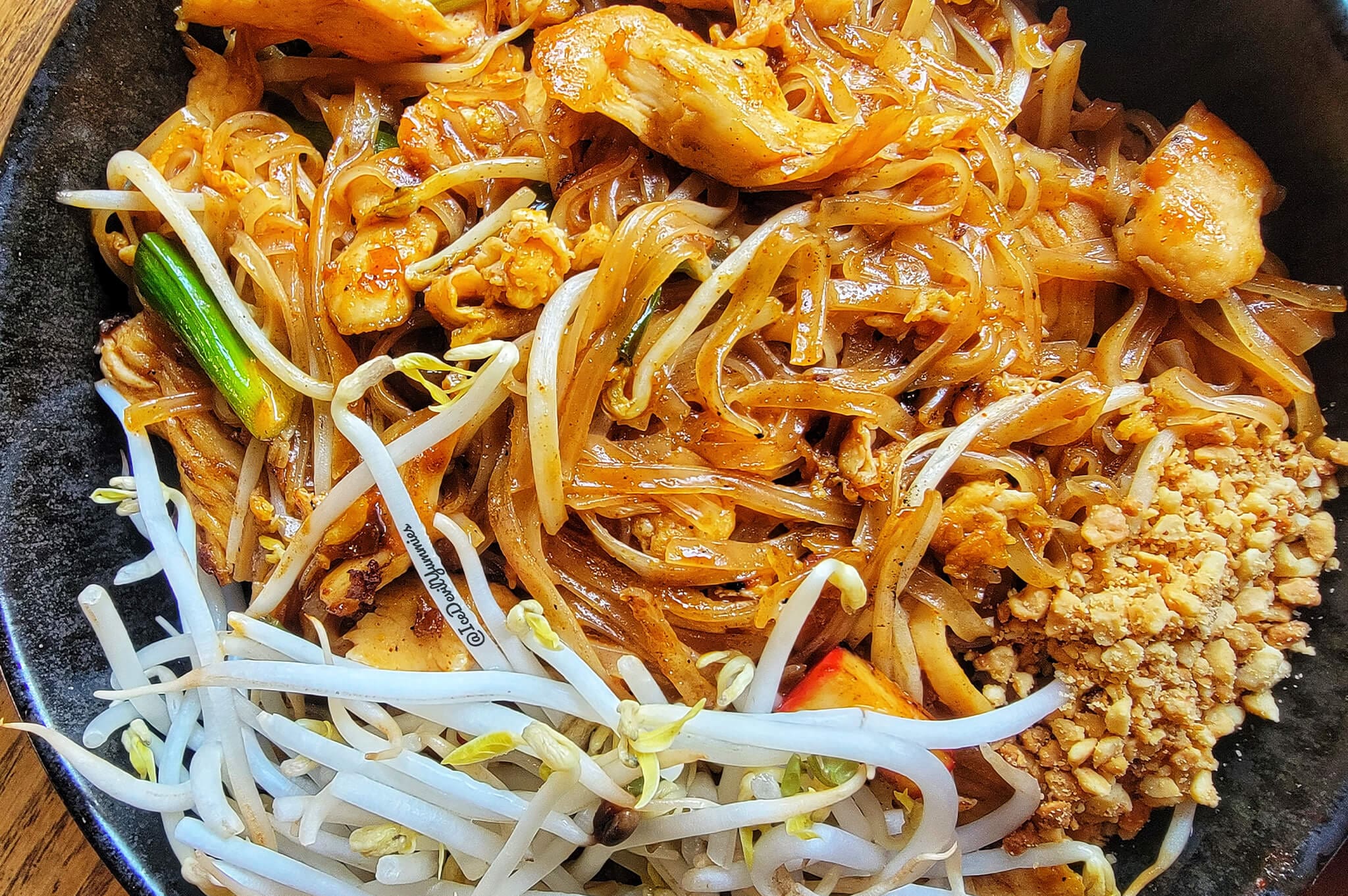 A bowl of Pad Thai featuring stir-fried rice noodles, bean sprouts, crushed peanuts, green onions, and pieces of chicken. The dish is garnished with a rich, savory sauce, enhancing its appetizing appearance.