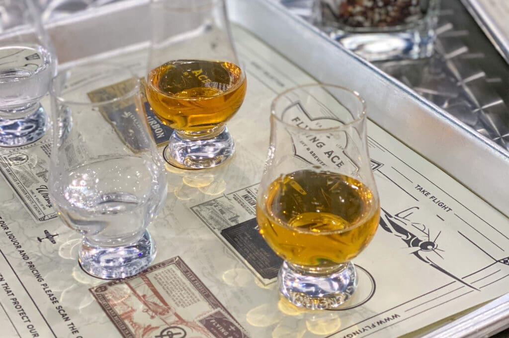 Four whiskey glasses, two filled with amber liquid, are placed on a metal tray with vintage-themed paper and airplane illustrations beneath them.