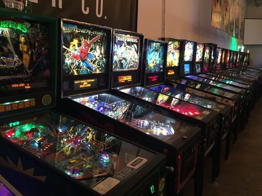 A row of colorful pinball machines is lined up in a dimly lit arcade. Each machine features vibrant artwork, including themes like superheroes and movies. The arcade is bustling with bright lights and a nostalgic atmosphere.