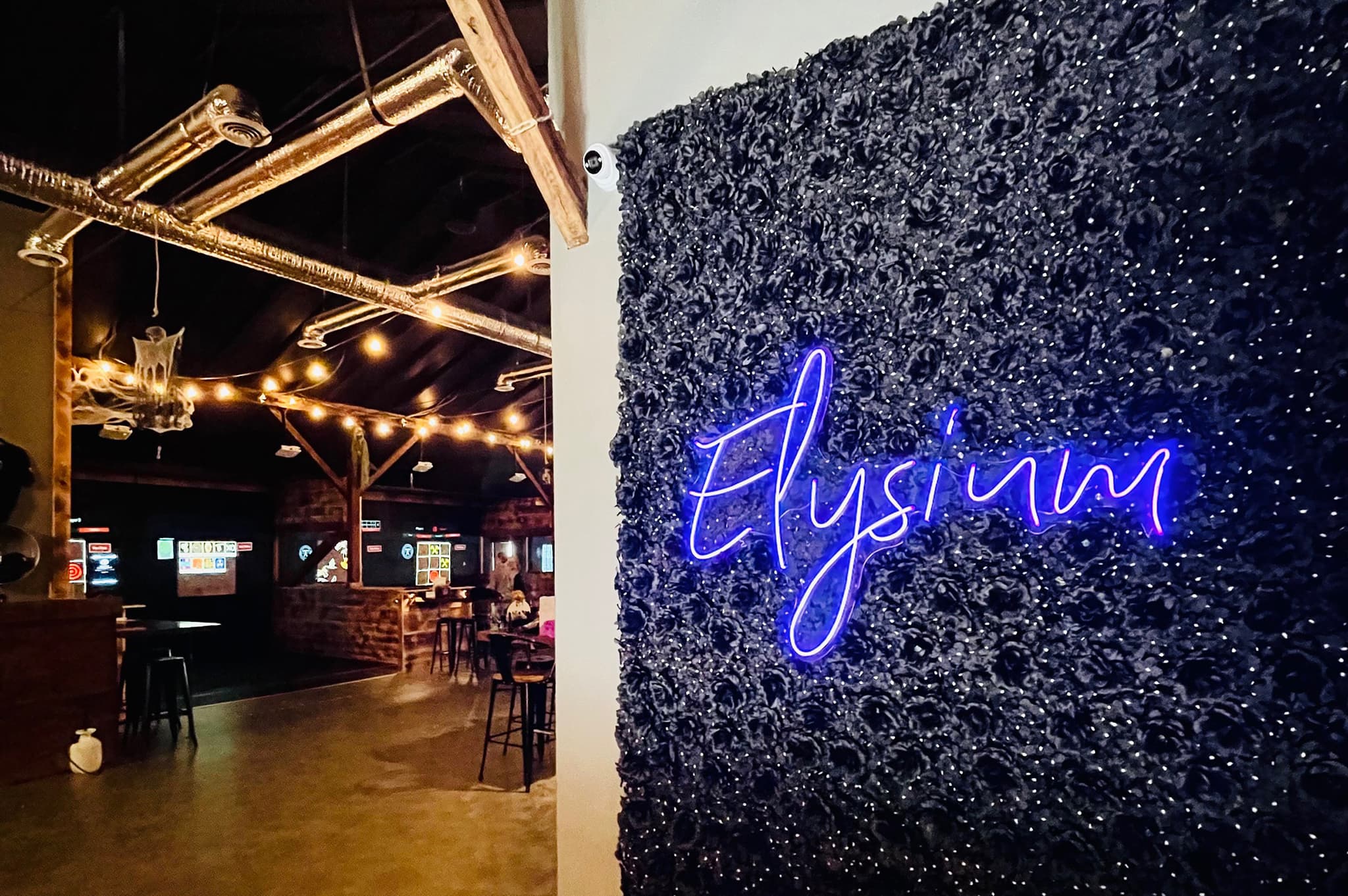 Neon sign reading Elysium on a textured black wall inside a dimly lit venue with string lights and exposed ductwork. High tables and stools are visible, creating a cozy atmosphere.