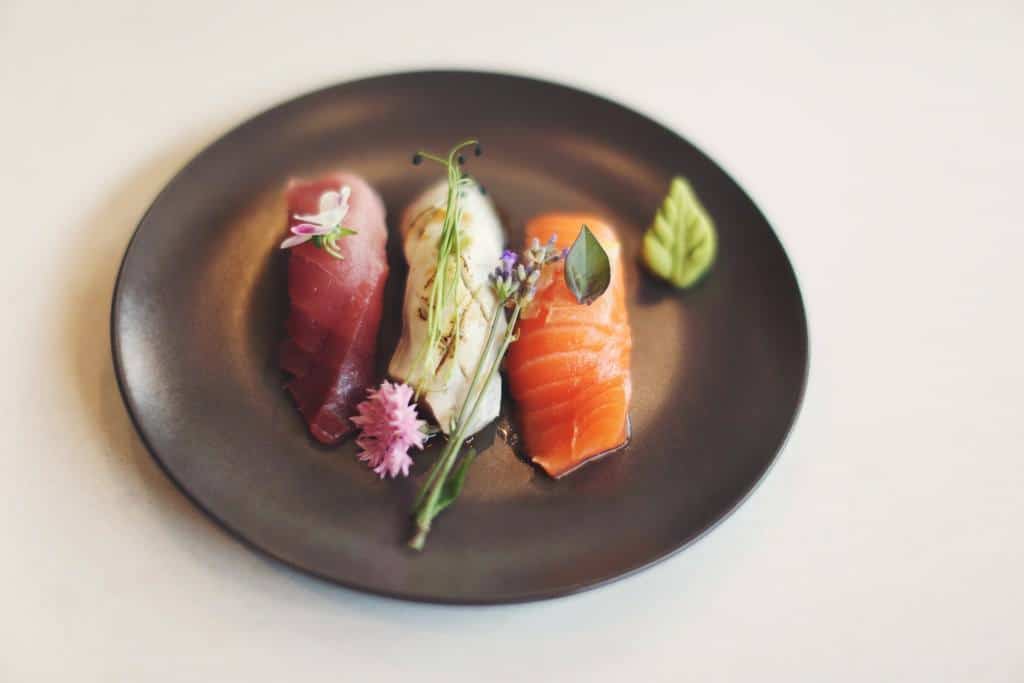 A dark plate holds three pieces of sushi: tuna, white fish, and salmon nigiri, garnished with small pink and purple flowers and a green leaf. The background is a softly blurred light surface.