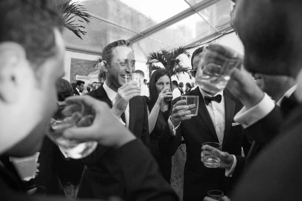 A group of people in formal attire, including tuxedos, are gathered under a tent raising glasses in celebration. The atmosphere is joyful, with palm plants visible in the background. Its a black and white photograph.