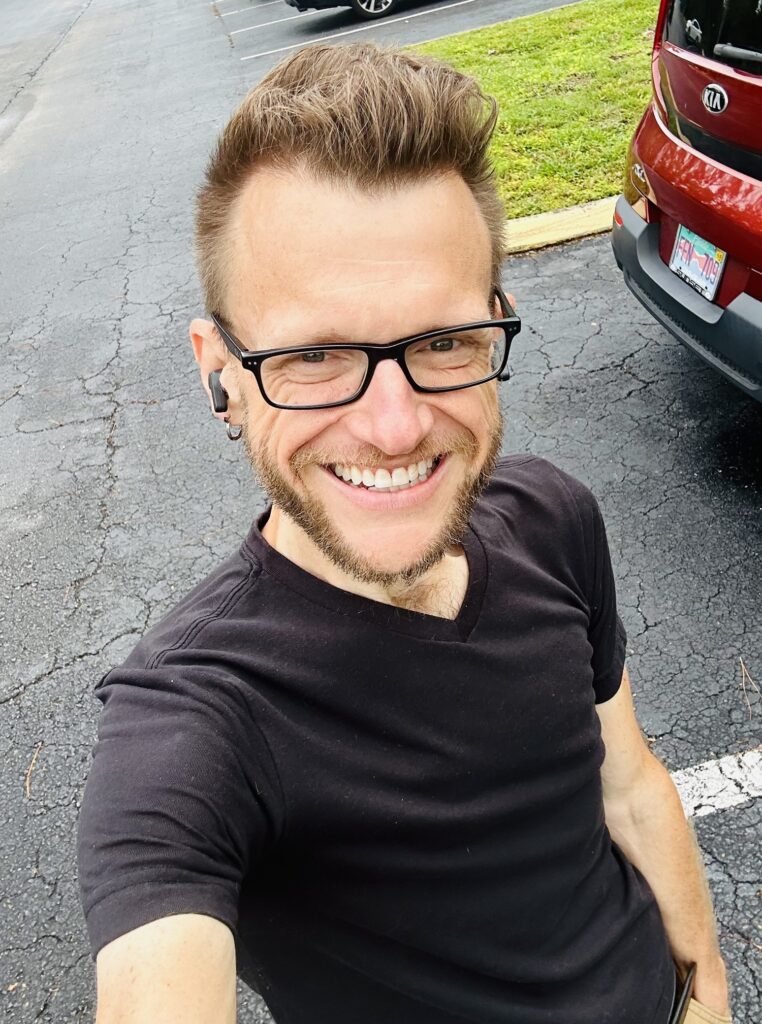 A smiling person with short hair, glasses, and a black t-shirt takes a selfie outdoors in a parking lot, with a red car and grass visible in the background.
