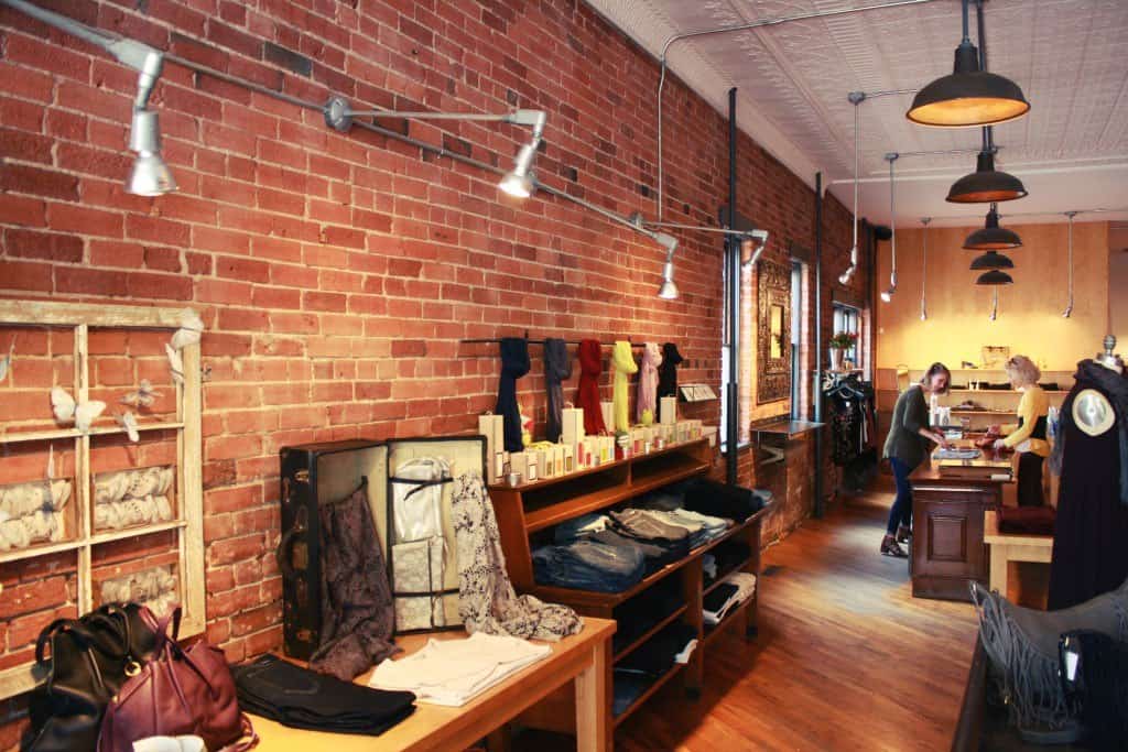 Cozy boutique with brick walls and wooden floors. Clothing and scarves are displayed on shelves and tables. Two people are interacting at a wooden counter on the right side, with hanging lights adding warmth to the space.