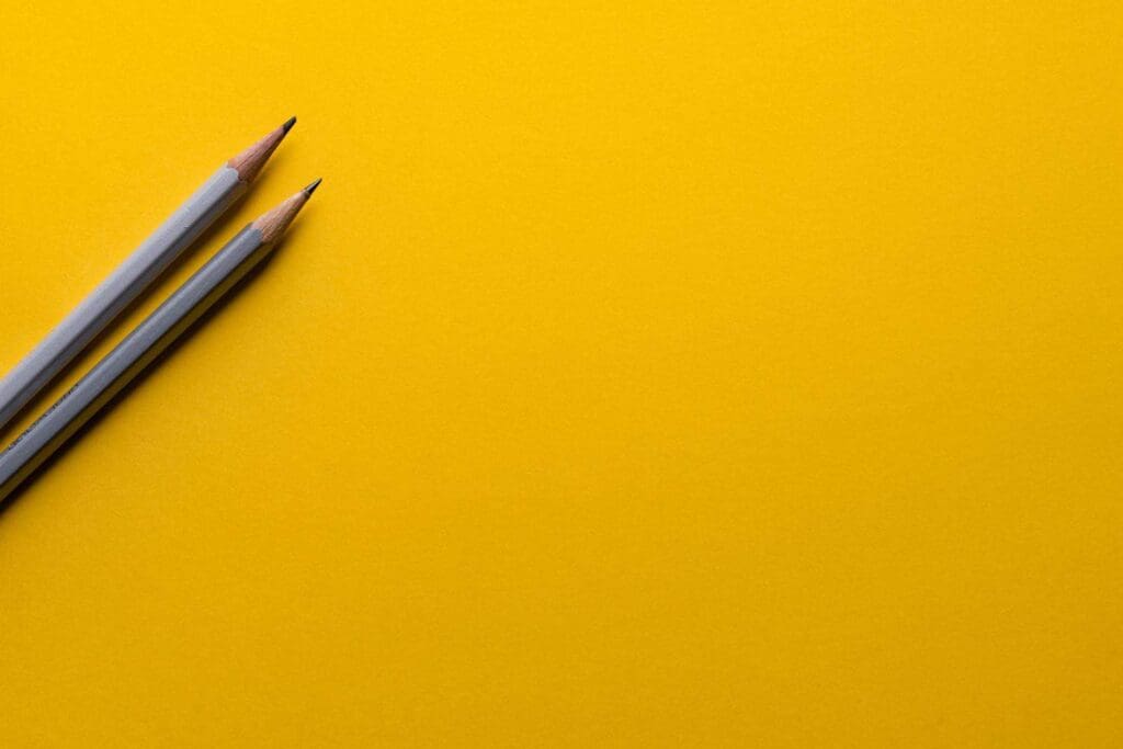 Two sharpened pencils with gray bodies lie diagonally on the left side of a bright yellow background. The background is smooth and untextured, creating a vibrant contrast with the pencils.