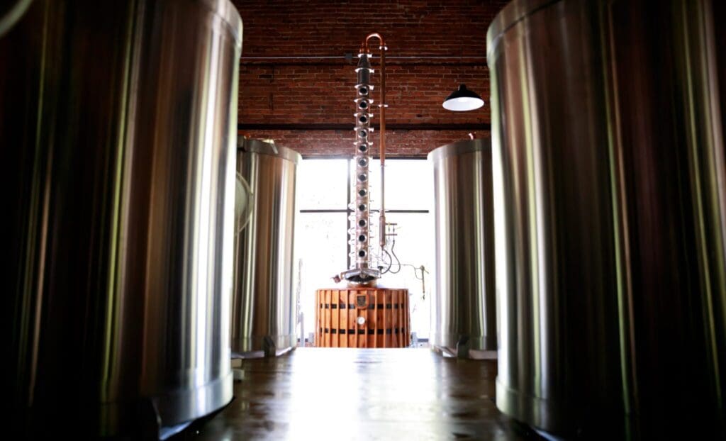 View of a distillery interior with large metal tanks on either side and a column still in the center. The space is well-lit with natural light coming through a window in the background. The ceiling is made of brick with an industrial lamp hanging.