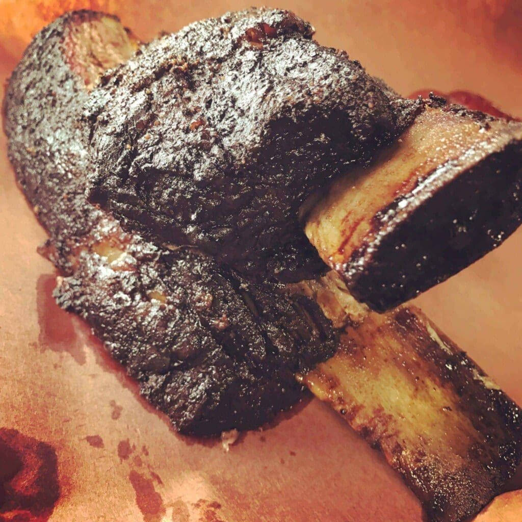 A close-up of a smoked beef rib resting on a piece of brown parchment paper. The rib is dark and charred, with visible marbling on the meat and bone. The texture appears tender and juicy.