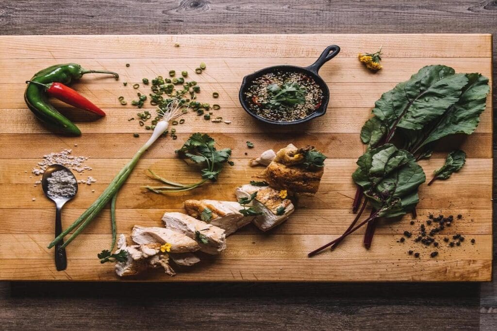 A wooden cutting board with chopped cooked chicken, beet greens, peppers, a small skillet with seeds and herbs, sliced green onion, and scattered spices, including chili flakes and coriander leaves. A spoon holds coarse salt.