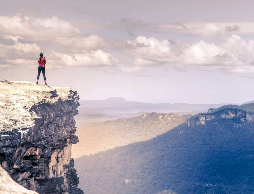 A person in a red jacket stands at the edge of a rocky cliff, gazing out over a vast landscape of rolling hills and forests under a partly cloudy sky. The scene conveys a sense of adventure and vastness.