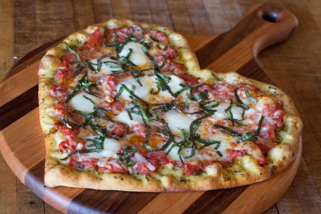 A heart-shaped pizza topped with mozzarella, tomato sauce, fresh basil, and bits of meat, placed on a wooden serving board. The pizza crust is golden brown and rustic, resting on a wooden surface.