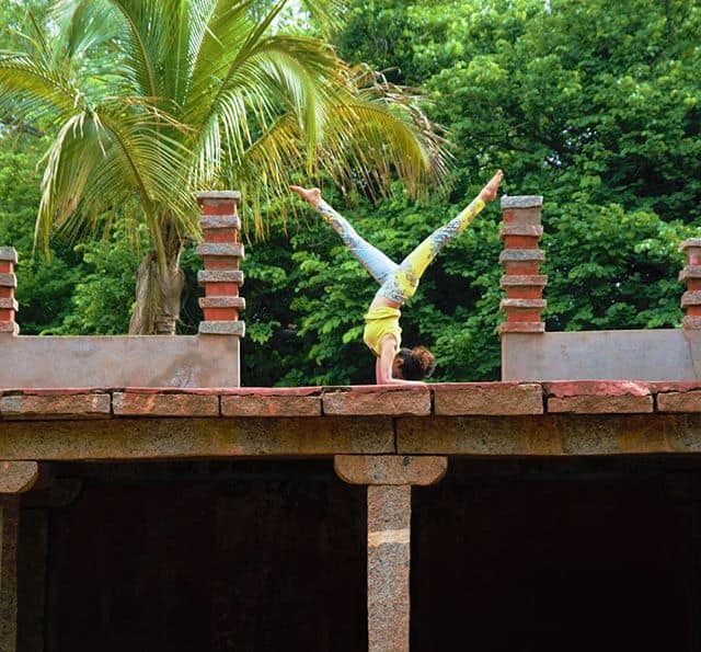 Person performing a handstand on a stone structure, with legs in a split position. A tall palm tree and lush greenery are in the background, under a clear sky.