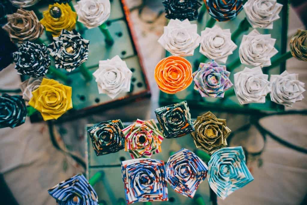 A collection of colorful, intricately folded paper roses displayed vertically, viewed from above. Each rose features unique patterns and colors, including white, orange, blue, and multicolored designs.