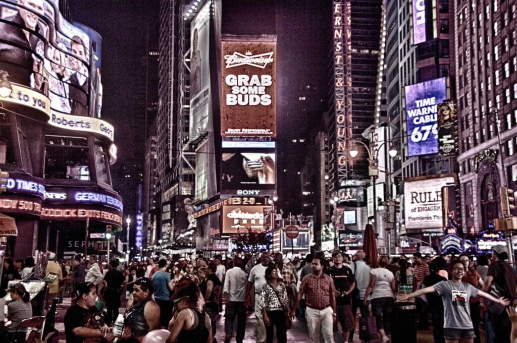 A vibrant, bustling scene of Times Square in New York City at night, filled with a crowd of pedestrians. Bright neon lights and large digital billboards illuminate the area, advertising various brands and attractions.