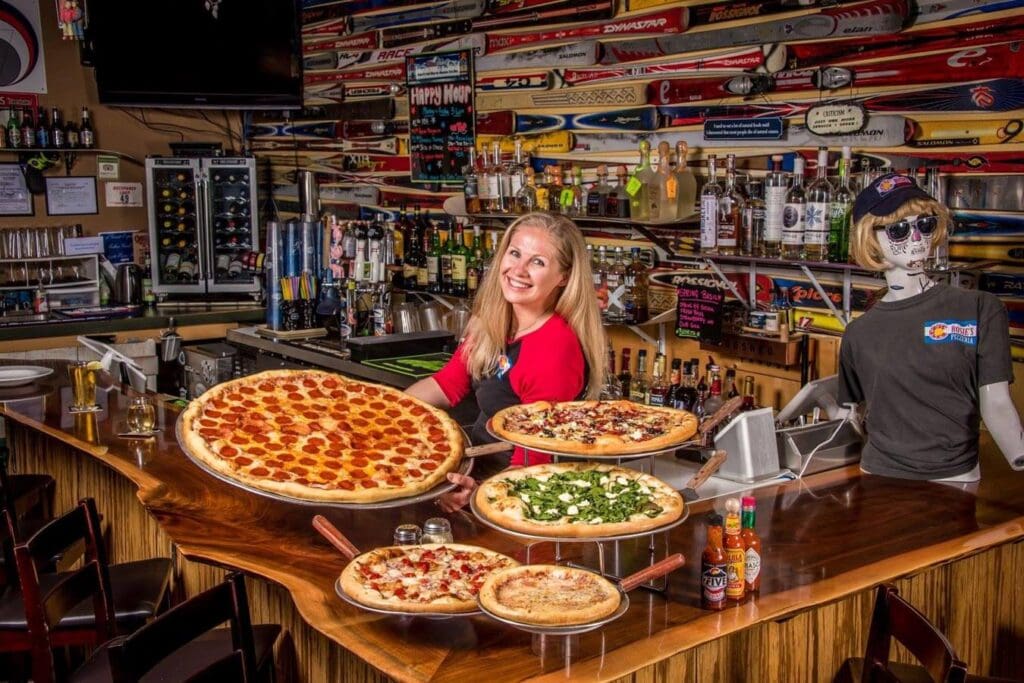 A woman smiles behind a bar counter displaying five pizzas of various toppings and sizes. The background features shelves with small boats and a mannequin with sunglasses and a cap. The setting is vibrant and colorful.